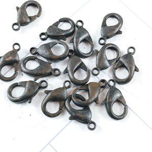 May include: A collection of antique bronze-colored lobster claw clasps. These metal clasps are designed for jewelry making and crafting. The clasps are scattered on a white surface, showcasing their detailed design and finish.