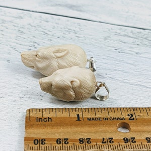 May include: Two cream-colored bear pendant charms with silver-colored bails. The pendants are carved with detailed bear faces. A wooden ruler with inch measurements is in the foreground.