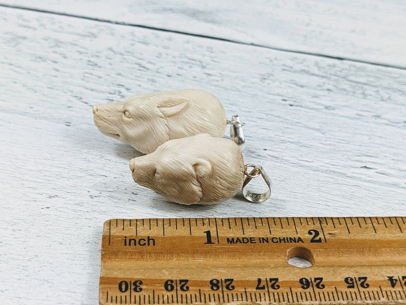 May include: Two cream-colored bear pendant charms with silver-colored bails. The pendants are carved with detailed bear faces. A wooden ruler with inch measurements is in the foreground.