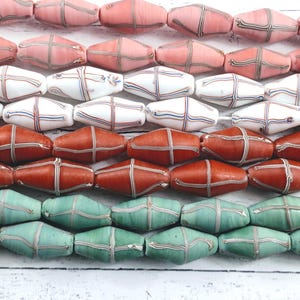 May include: Rows of colorful, oblong beads in shades of pink, white, red, and teal. Each bead features a cross-hatch design in contrasting colors. The beads are arranged horizontally, showcasing their unique patterns and hues.