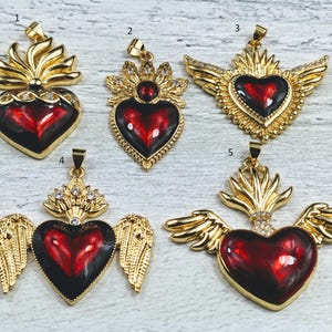 May include: Five gold-tone pendants with red heart-shaped glass insets. Each pendant features a different design, including wings, flames, and a crown. The pendants are all on a white background.