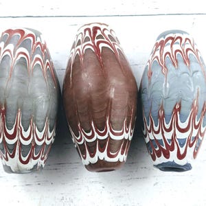 May include: Three decorative glass beads with a unique pattern. The beads are oval-shaped, with a base colour of grey, brown, and blue. Each bead features a wavy design in white and red, creating a striking visual effect. These beads are ideal for jewellery making or decorative crafts.
