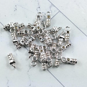 May include: A collection of small, silver-colored jewelry components. Each component has a cylindrical body with decorative details and a small loop at one end. Some pieces have clear, round embellishments.
