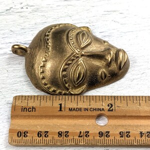 May include: A gold-toned metal pendant in the shape of a stylized human face. The face has detailed features and a textured surface. The pendant is approximately 2 inches long.