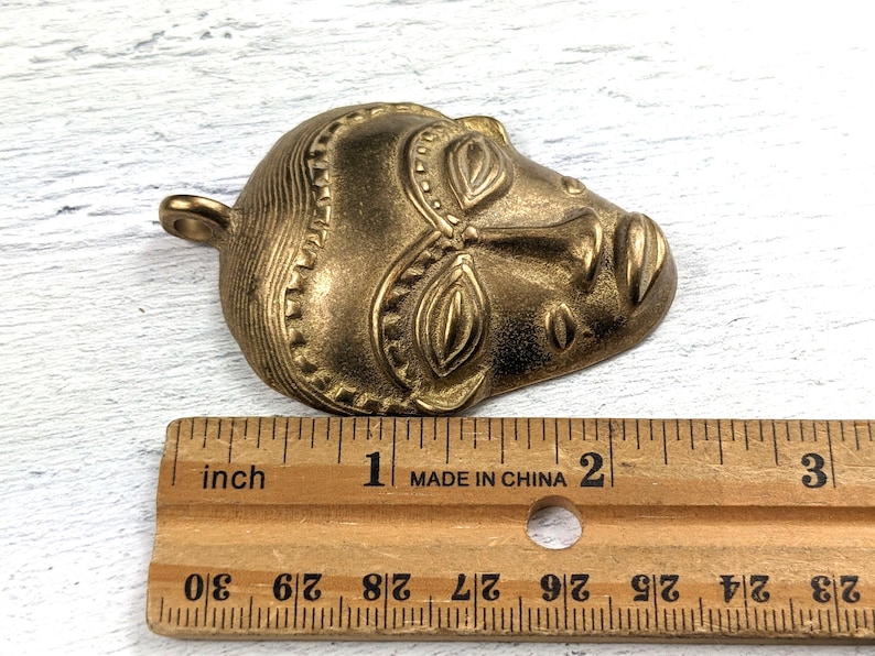 May include: A gold-toned metal pendant in the shape of a stylized human face. The face has detailed features and a textured surface. The pendant is approximately 2 inches long.