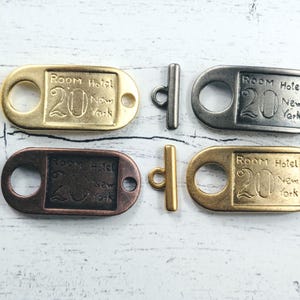 May include: Four metal toggle clasps in gold, bronze, and silver tones. Each clasp features a rectangular tag with the text "Room Hotel 20 New York". The clasps are for jewellery making or crafting.