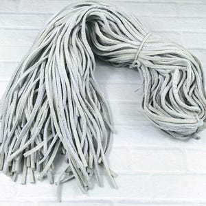 Puede incluir: Un manojo de cordón de cuero blanco, de aproximadamente 3 metros de largo, se muestra en una bobina suelta. El cordón está hecho de tiras finas y planas de cuero que están entrelazadas.