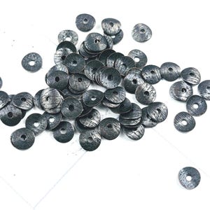 May include: A collection of dark gray, textured metal beads with a central hole. The beads are round and flat, scattered on a white surface. The beads have a distressed, weathered appearance.