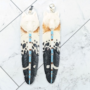 May include: Two carved feather pendants. Each pendant features a wolf or bear head at the top, with a feather design in cream, brown, black, and turquoise. The pendants are displayed on a white and gray tiled surface.