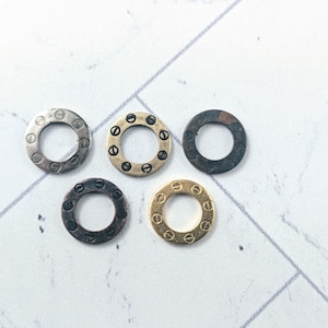 Puede incluir: Cinco anillos metálicos con diseño circular y pequeños tornillos en el borde. Los anillos son de color plata, oro y negro. Están dispuestos sobre una superficie blanca. Ideales para manualidades o joyería.