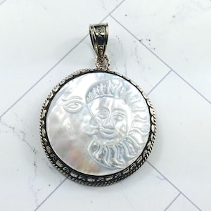 May include: A silver pendant with a white mother of pearl inlay depicting the sun and moon. The pendant is round and has a detailed silver border.
