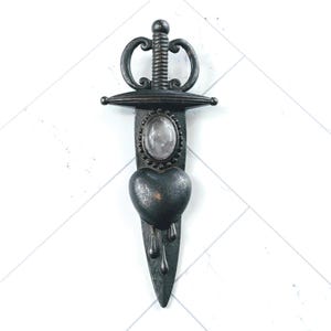 May include: A black metal dagger with a heart-shaped blade and a clear crystal in the center. The dagger is dripping with blood.