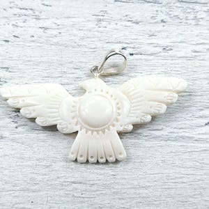 May include: An off-white pendant carved in the shape of a bird with wings spread wide. The center of the bird features a smooth circular detail surrounded by etched lines. A silver-toned metal bail is attached to the top of the bird's head.
