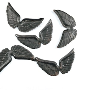 May include: A collection of five dark metallic wing-shaped ornaments, each featuring detailed feather textures and a curved design. These individual pieces are arranged in a scattered pattern, showcasing a weathered, dark bronze-toned finish.