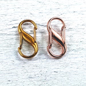 May include: Two S-shaped metal clasps, one gold and one rose gold, on a white wooden background.