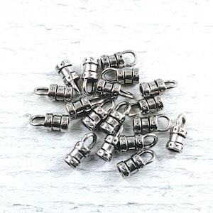 May include: A collection of silver-colored metal cord end crimps with a loop for attaching to jewelry. Each crimp has a cylindrical shape with decorative details and a small loop for attaching to a chain or other jewelry component.