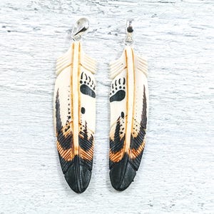 May include: Two feather pendants with a bear paw print design. The pendants are cream-colored with black, brown, and orange accents. Each feather has a silver-colored bail at the top for hanging.