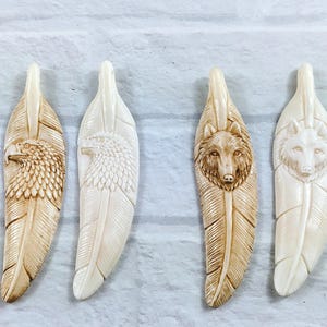 May include: Four carved feather pendants. Two feature eagle designs, and two feature wolf designs. The pendants are off-white and tan, with detailed carvings of the animals' faces and feather textures. Each pendant is approximately 5 inches long.
