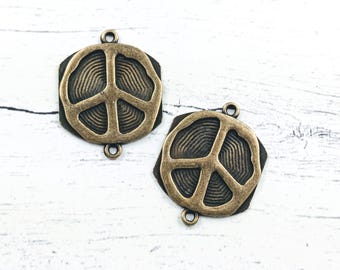 May include: Two bronze-colored peace sign charms. Each charm features a peace symbol within a textured, dark brown border. The charms have small loops for attaching to jewelry or other crafts. The background is a white, textured surface.