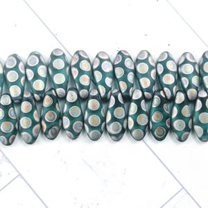 May include: Teal-colored glass beads with a polka dot pattern. The beads are oval-shaped with a flattened top and bottom. The polka dots are a mix of white, gray, and light orange. The beads are arranged in a row, suitable for jewelry making.