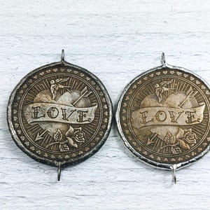 May include: Two silver-toned metal charms with a heart design, an arrow, and the word "LOVE" inscribed on a banner. Each charm has a small loop at the top for attaching to a necklace or bracelet.