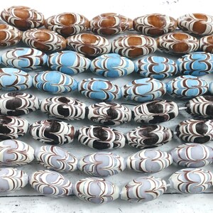 Puede incluir: Primer plano de cuentas ovaladas en varios colores. Las cuentas presentan un patrón marrón, blanco y azul. Algunas son marrones y blancas, otras azules y blancas, y otras negras y blancas. Las cuentas están ensartadas.