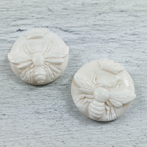 May include: Two white ceramic buttons with a raised design of a bee. The bee has detailed wings and a textured body.