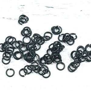 Puede incluir: Un conjunto de pequeños anillos de salto negros mate esparcidos sobre una superficie blanca. Estos anillos abiertos están destinados a la fabricación de joyas u otras manualidades. La imagen muestra una vista de cerca de los anillos, destacando su forma circular y color oscuro.