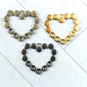 May include: Three heart-shaped metal embellishments in antique brass, gold, and silver tones. Each heart is composed of a series of small, round elements, some with holes, arranged in a heart shape. The background is a white, wood-like surface.