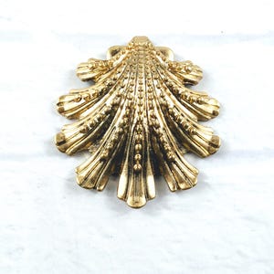 May include: A gold-toned metal brooch shaped like a scallop shell. The surface features a series of radiating ridges and raised beaded textures that follow the natural fan-like pattern of the shell, creating a metallic finish with dark highlights.