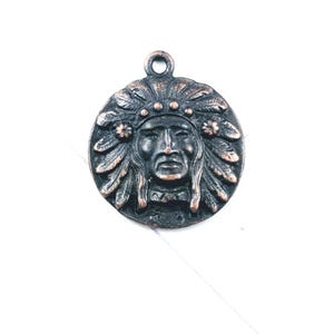 May include: A round, bronze-colored pendant featuring a detailed relief of a Native American chief in a feathered headdress. The pendant has a loop at the top for hanging and is set against a white background. The design is intricate and the metal has a slightly aged appearance.