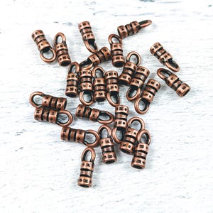 May include: A group of 20 copper-toned metal crimp end caps. The caps are cylindrical with a small loop at one end. The caps are scattered on a white surface.