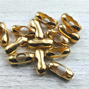 以下が含まれることがあります： 金色の金属製のチェーンコネクターのグループ。コネクターは楕円形で、中央に穴が開いています。白い表面に積み重ねられています。