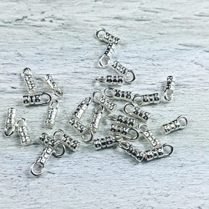 May include: A group of silver-toned metal crimp beads with a small loop on each end. The beads are arranged in a random pattern on a light wood surface.