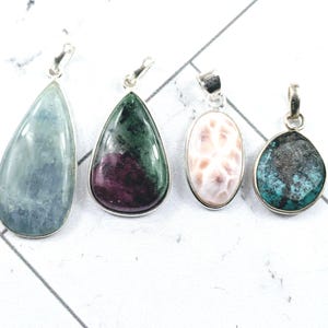 May include: Four gemstone pendants with silver-colored bails. The stones are teardrop, oval, and irregular shapes, in shades of blue, green, pink, and turquoise. The pendants are displayed on a white surface.