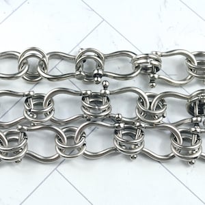 May include: Close-up of three rows of silver-colored metal chain links. The links are intricately designed with a series of interlocking circles and bars, creating a decorative pattern. The chain is displayed against a white and light gray background.