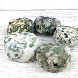 May include: Five moss agate stones with a white and green mottled pattern. The stones are smooth and polished, and each has a hole drilled through it.