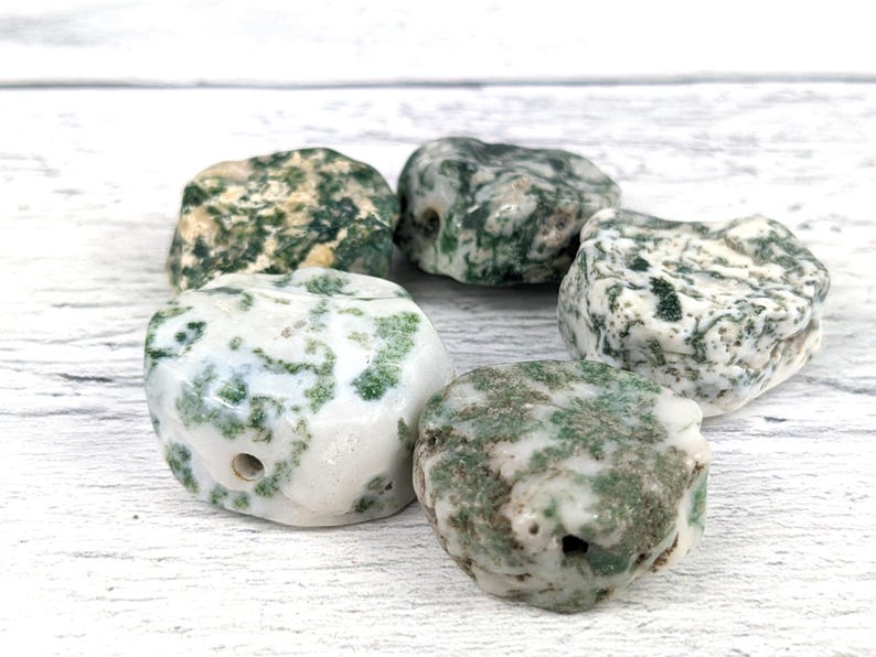 May include: Five moss agate stones with a white and green mottled pattern. The stones are smooth and polished, and each has a hole drilled through it.