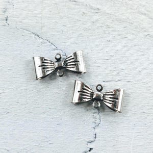 May include: Two silver-coloured bow-shaped charms with a textured finish. Each charm has a loop at the top and bottom for attaching to jewellery or other crafts. The charms are displayed on a light blue and white textured surface.