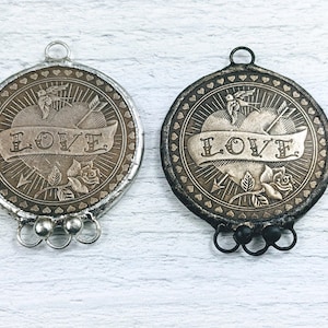 Puede incluir: Dos colgantes de metal plateado y negro con un diseño de corazón y la palabra "LOVE" en una pancarta. Cada colgante tiene un diseño de pájaro y flecha y está unido a una cadena con tres anillos.