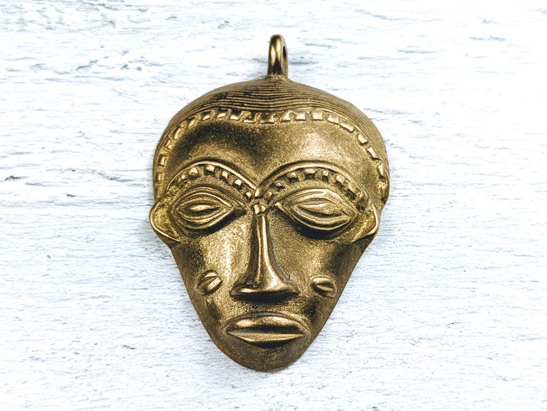 May include: A gold-colored metal pendant in the shape of a stylized human face. The face has detailed features, including eyes, a nose, and a mouth.