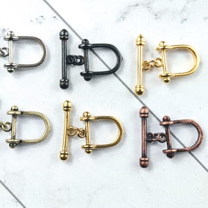 May include: A collection of six metal toggle clasps in various finishes: silver, black, gold, antique brass, and copper. Each clasp features a T-bar and a D-shaped loop, ideal for jewelry making or craft projects.