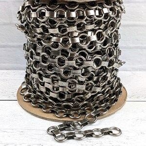 May include: A spool of gunmetal-colored chain featuring a repeating pattern of circular links connected by flat, rectangular metal segments. The chain is wound tightly around a circular cardboard core, with a single strand extending from the base.