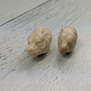 May include: Two small, beige bear head carvings. The bear heads are detailed with visible fur and facial features. They are positioned on a light-colored, textured wooden surface.