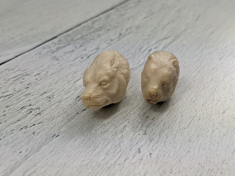 May include: Two small, beige bear head carvings. The bear heads are detailed with visible fur and facial features. They are positioned on a light-colored, textured wooden surface.