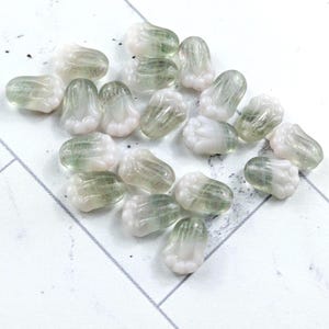 May include: A collection of translucent, flower-shaped beads. Each bead has a white base with a green-tinted centre, resembling a delicate floral design. The beads are scattered on a white surface with a subtle geometric pattern.