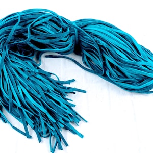 Puede incluir: Un manojo de tiras de cuero planas de color cerceta dispuestas en una formación curva y suelta. Las tiras son uniformes en ancho y textura, con un acabado mate y un tono azul verdoso vibrante y constante en toda la colección.