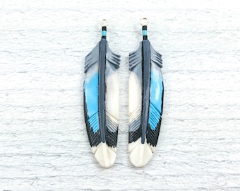 May include: Two feather-shaped pendants. Each pendant features a design with gray, light blue, black, and white colors. The top of each feather has a gold-colored loop for hanging. The feathers are carved with intricate details.