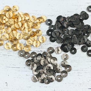 May include: A collection of gold, silver, and black metal round disc-shaped embellishments. The embellishments are scattered on a white wooden surface.