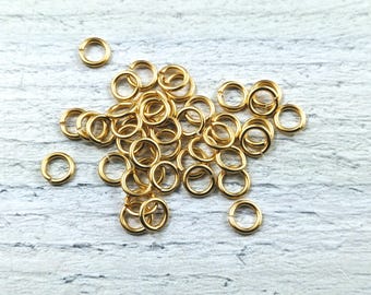 May include: A pile of gold jump rings, a type of jewelry finding, on a white wooden surface.